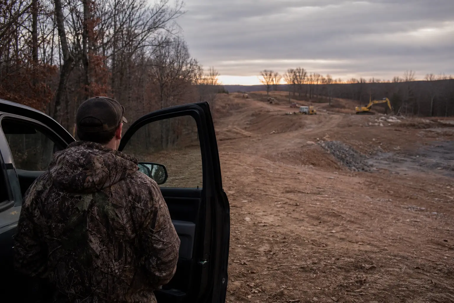 A person in camouflage sits in the open doorway of a pickup truck, looking down a muddy rural road