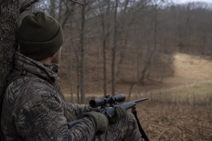 Camouflaged person in a knit cap aiming a rifle with a scope while seated in a wooded field