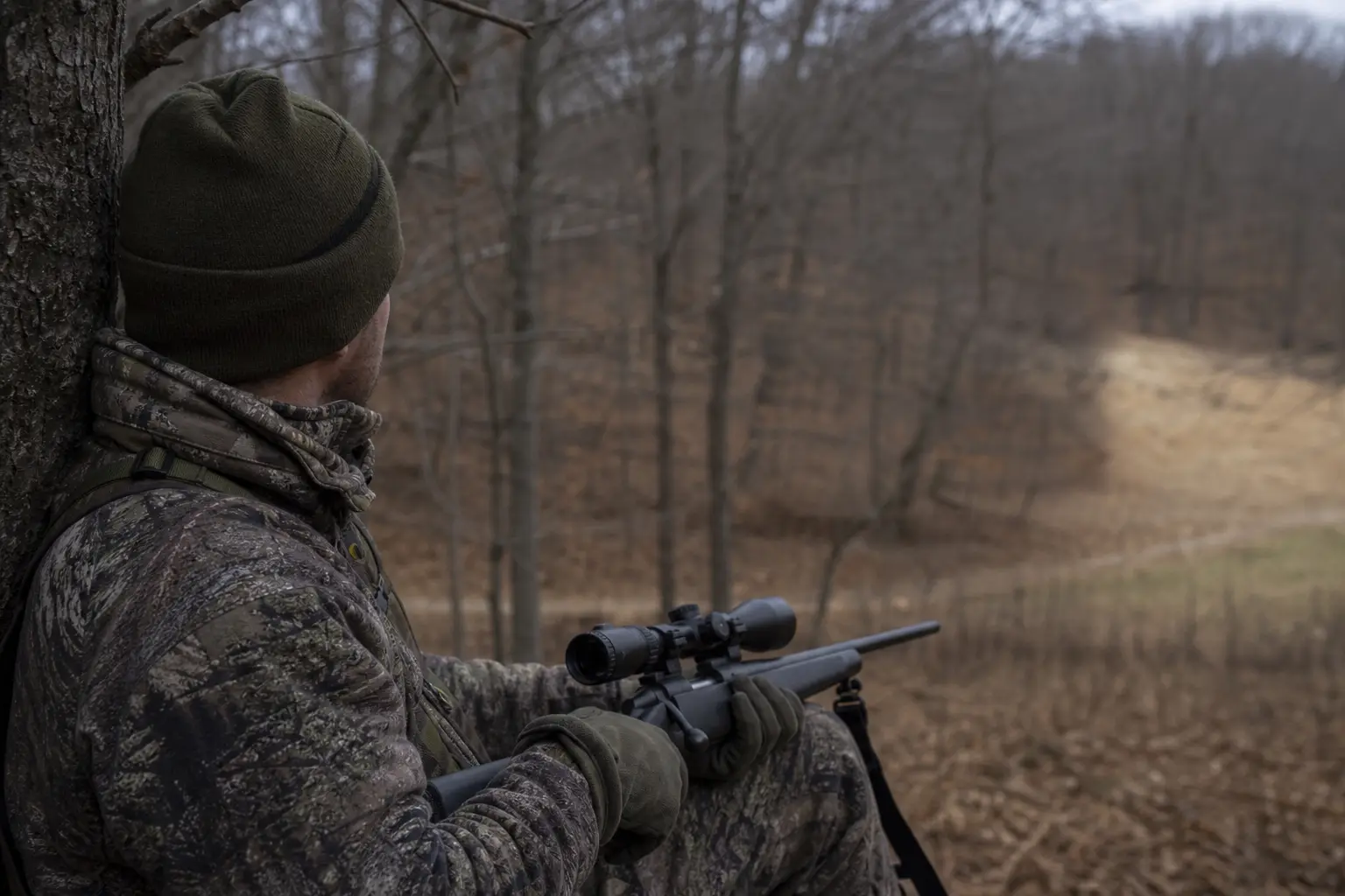 Camouflaged person in a knit cap aiming a rifle with a scope while seated in a wooded field