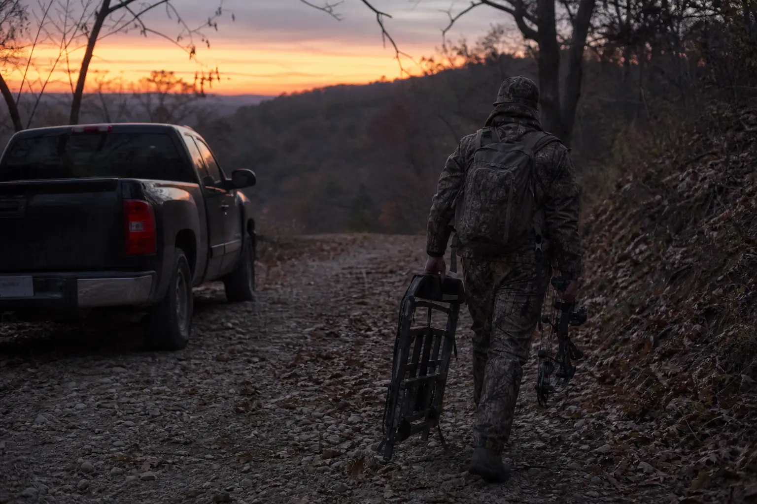 Camouflaged person in helmet pushes a bicycle along a rocky road beside a parked pickup at sunset