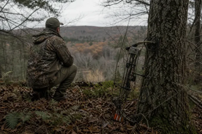 A camouflaged person crouches in a forest with a tripod-mounted device beside a tree, looking toward distant hills