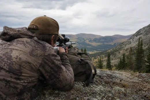 Two people in camouflage clothing aim binoculars and a scope from a rocky hillside overlooking a mountain valley