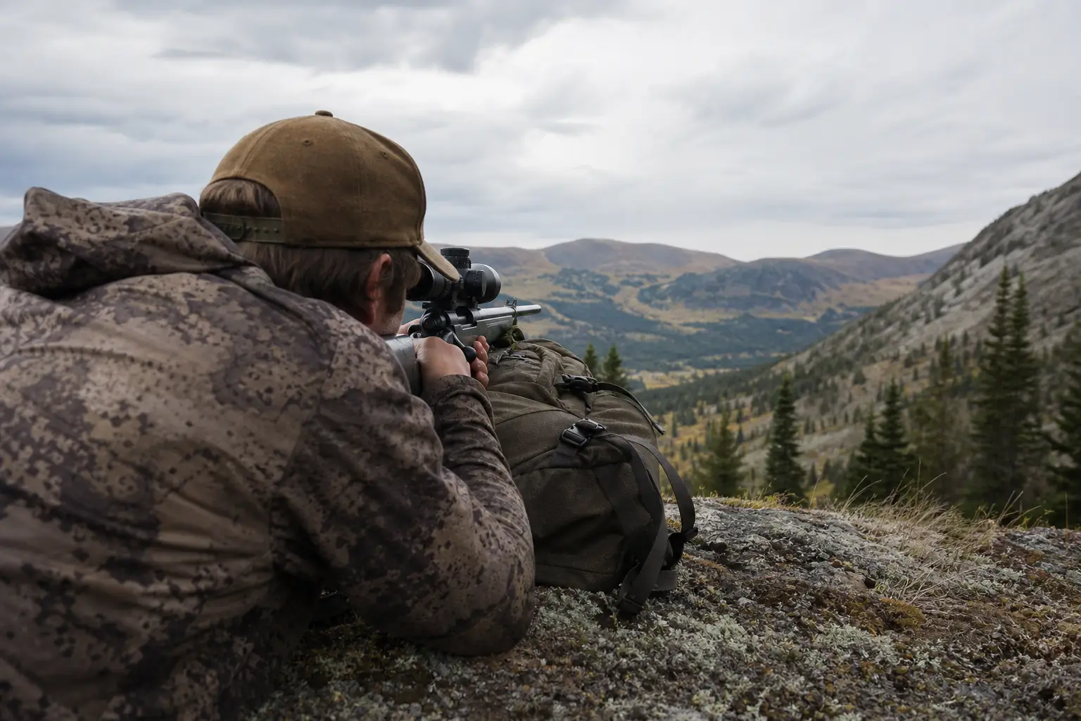 Two people in camouflage clothing aim binoculars and a scope from a rocky hillside overlooking a mountain valley