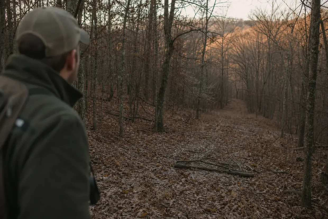 Person in a cap and jacket stands at left, looking down a leaf-covered dirt path through bare trees