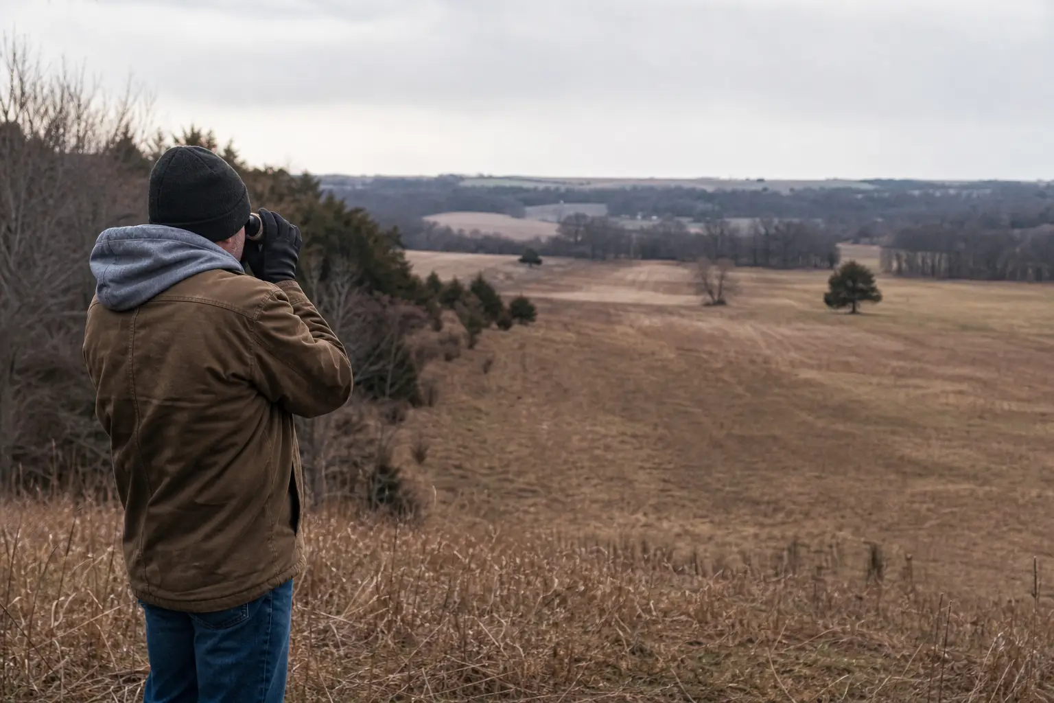 Person standing outdoors with binoculars, looking across a wide rural field with trees and low hills in the distance