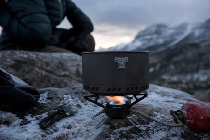 Camping stove with blue-orange flame on rocky ground, person in dark jacket kneeling nearby against snowy mountains