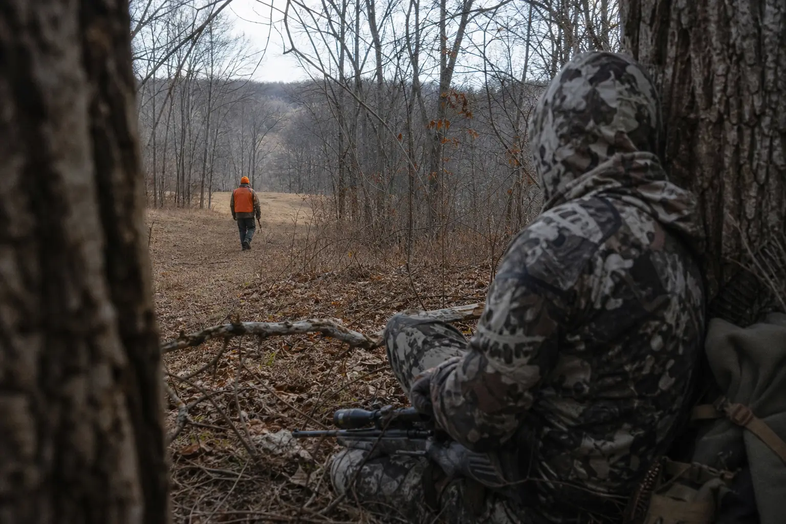 Two people in camouflaged clothing crouch in a wooded area with a trail of branches and a distant hiker