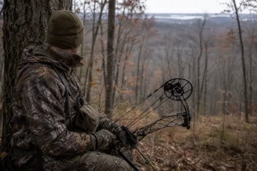Camouflaged person crouches behind a tree, holding a compound bow with a black wheel-like sight in a wooded area
