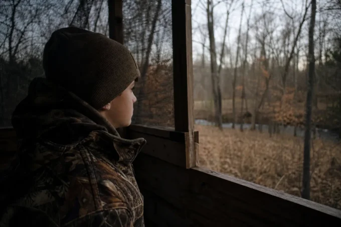 Person in a dark knit hat and camouflage jacket sits by a window, looking out toward leafless trees