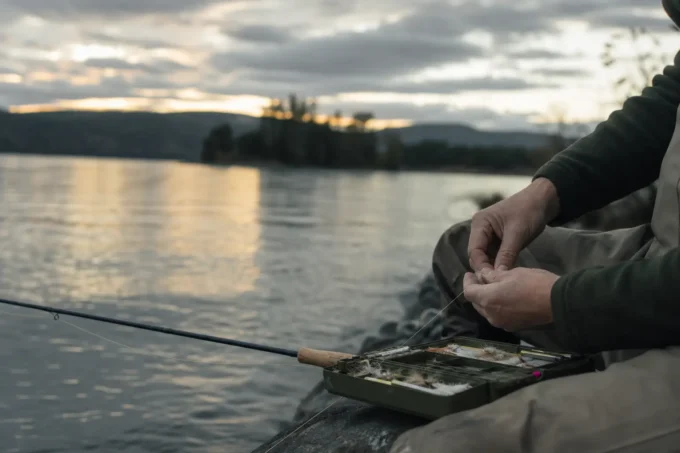 Two hands by a boat seat near a fishing tackle tray, on a calm lake at sunset with hills in distance