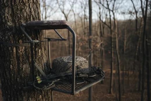 Camo crossbow mounted on a metal stand attached to a tree trunk, with a wooded background behind it