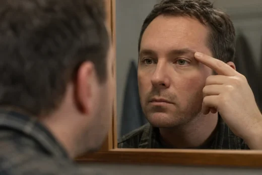 Man looks into a mirror, raising his hand to his forehead beside his eye