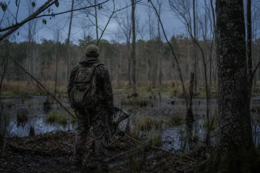 Person in camouflage with a backpack stands by a pond in a wooded area, holding a bow-like device