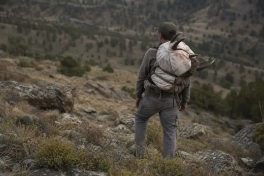 Person stands on a rocky hillside, wearing a backpack and camouflage clothing, with a mountainous landscape in the background