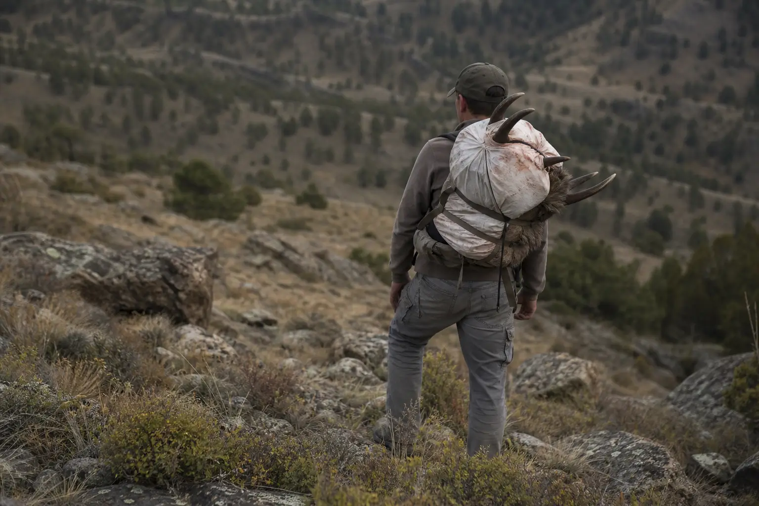 Person stands on a rocky hillside, wearing a backpack and camouflage clothing, with a mountainous landscape in the background