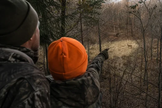 Two people in camouflage and dark clothing point toward a wooded trail, with an orange cap in the foreground