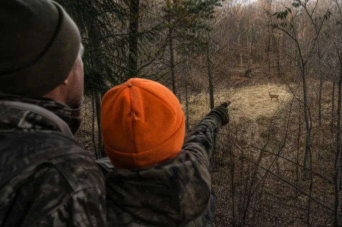 Two people in camouflage and dark clothing point toward a wooded trail, with an orange cap in the foreground