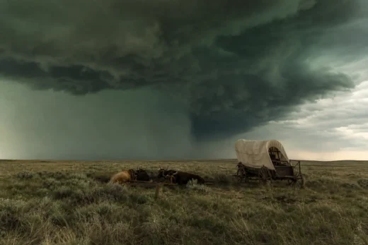 Wagon with canvas cover and horses lying in grass under large dark storm clouds across a flat landscape