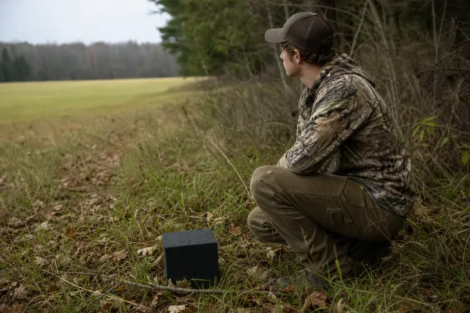 Person in camouflage clothing and cap kneels in grassy field, holding a handheld device near a dark box