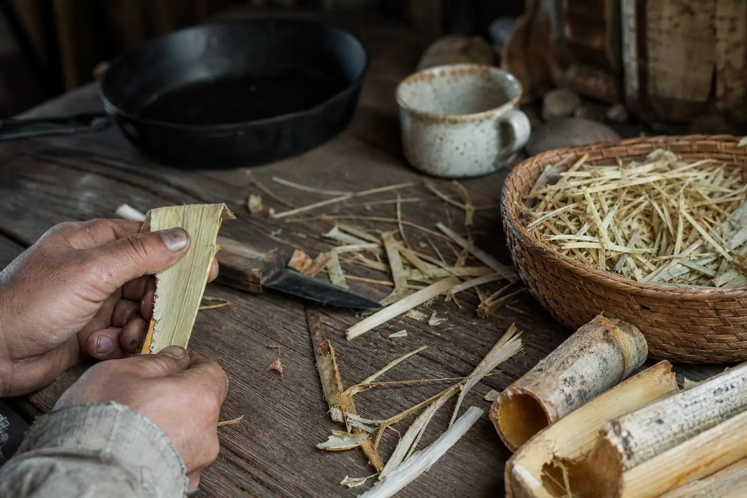Hands split a piece of plant stalk on a wooden table, surrounded by cut bamboo, straw, and metal cups