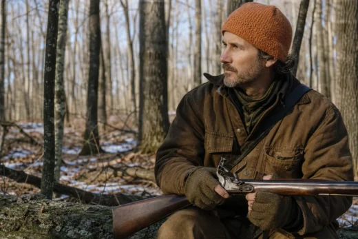 A man in a brown knit cap and jacket sits outdoors in a leafless forest holding a long rifle