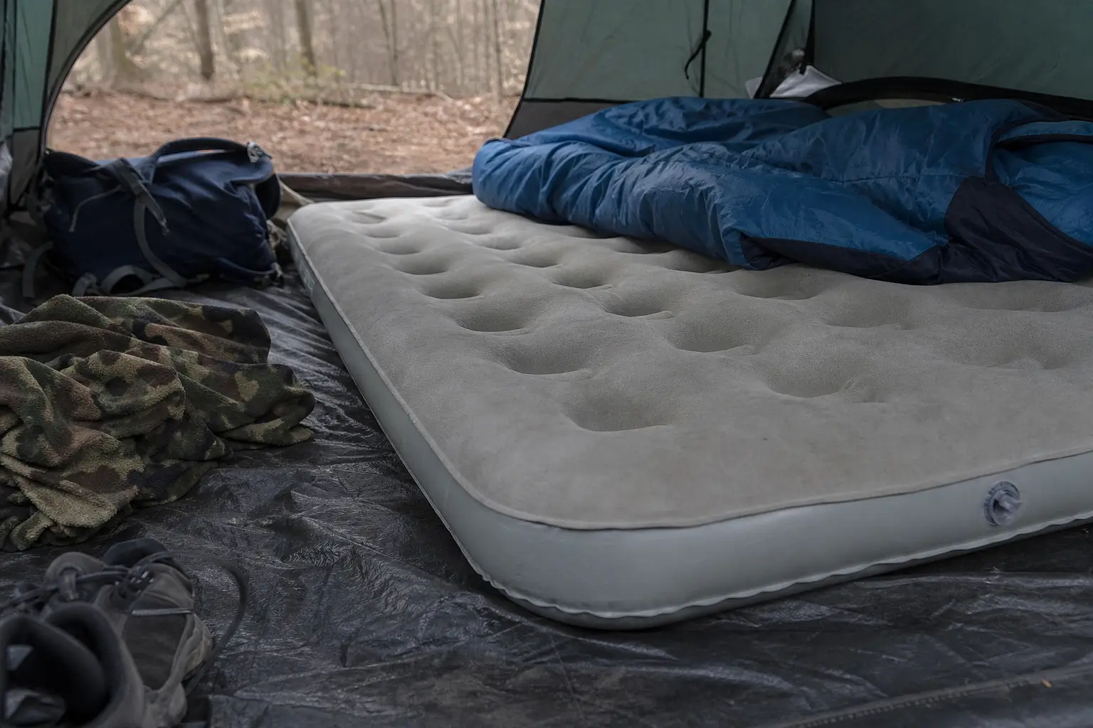Open tent with gray air mattress, blue sleeping pad, and folded camouflage blankets on the floor