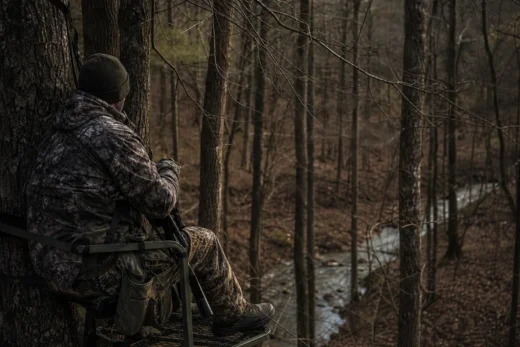 Camouflaged person in a forest, leaning behind a tree while holding a rifle and looking outward