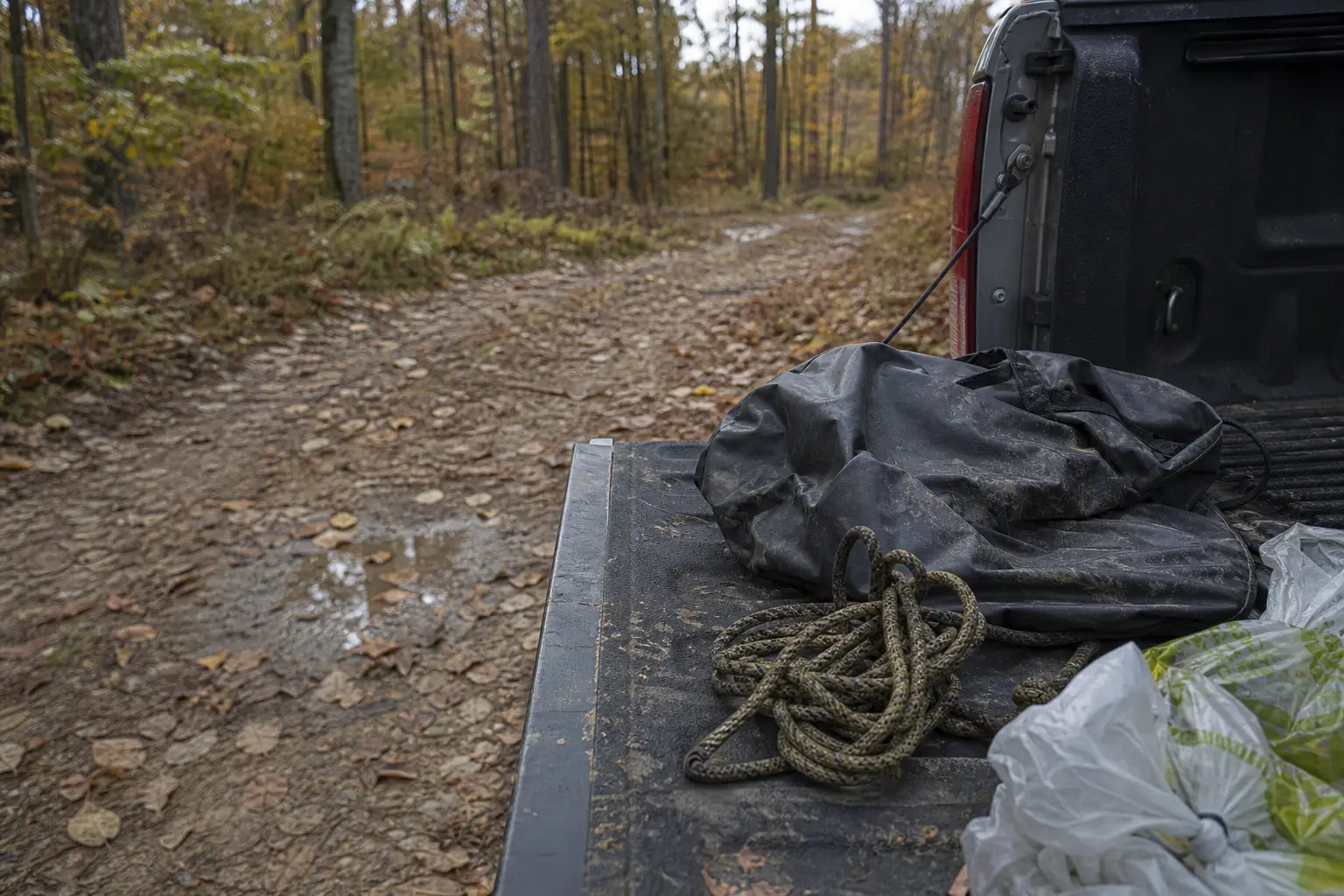Truck bed at roadside with black tarp, coiled rope, and a plastic bag, surrounded by an autumn forest trail