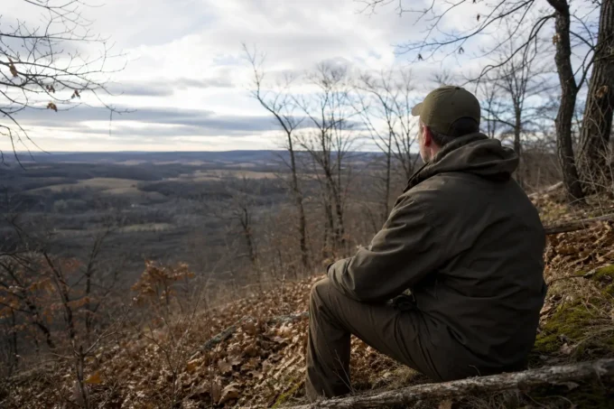Person in an olive jacket and cap crouches on a rocky hillside, looking over a distant valley under cloudy skies