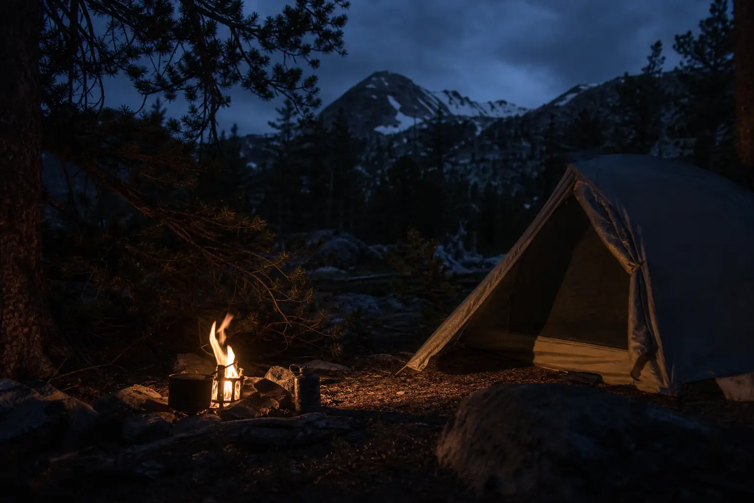 Tent beside a small campfire with flames glowing on rocky ground; pine trees and snow-covered mountains in the distance