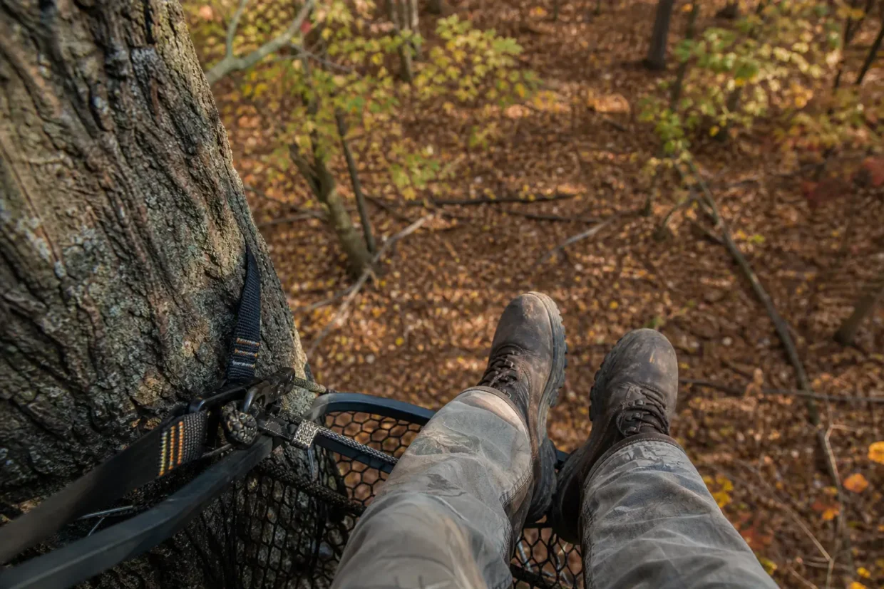 Camouflage netting tied to a tree with a bow-like device; person’s boots and pant legs rest in a wooded area