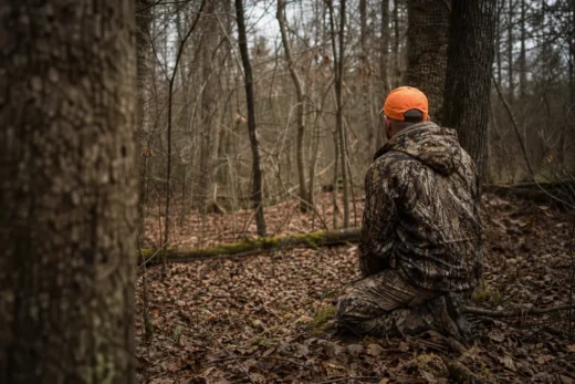 Person crouching in a leaf-covered forest path, wearing a camouflage jacket and orange cap, seen from behind