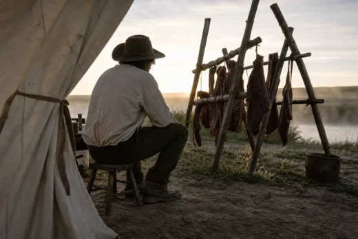 Man in hat kneeling by a table under a tent, with hanging meats on metal racks near a river at sunset